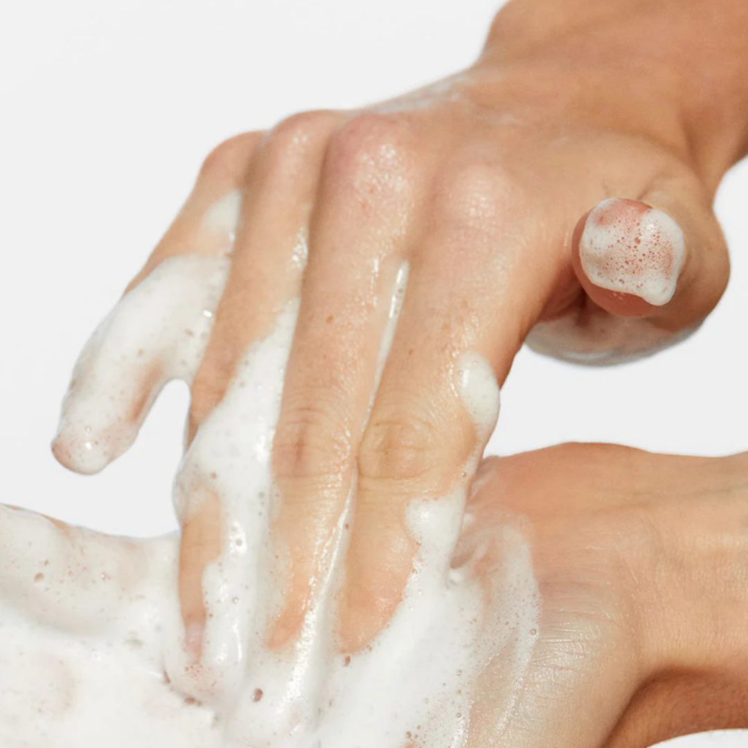 Close-up of hands covered in white foamy cleanser lather on light background